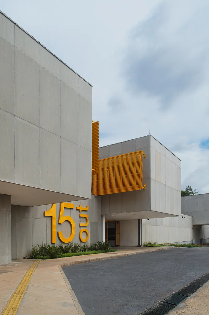 Street entrance with concrete facade, yellow metal louvers, and large dimensional numerals beside planted beds under cloudy sky