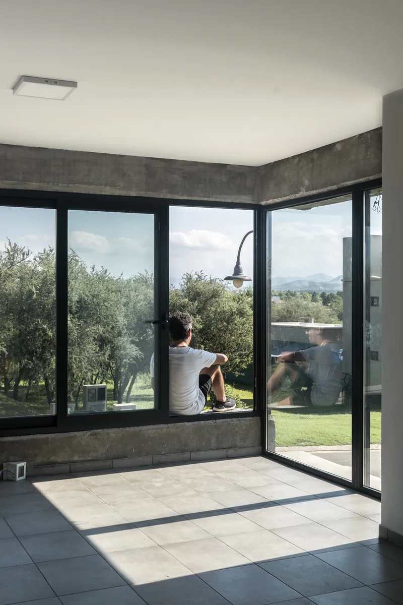 Interior corner with floor-to-ceiling glazing, concrete beam soffit, and person seated on window sill
