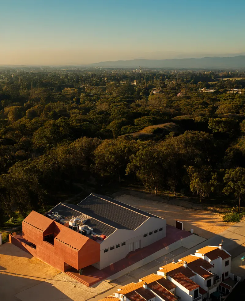 Aerial view of terracotta and white gabled volumes with corrugated metal roofs in forested landscape at sunset