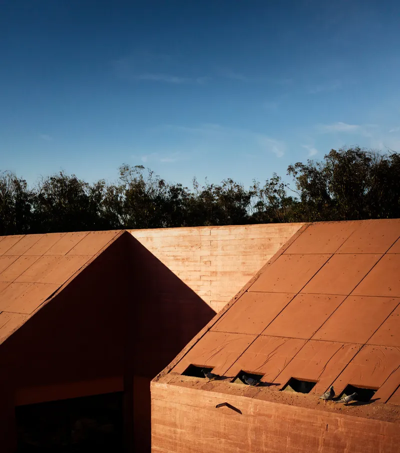 Close-up of terracotta brick roofline junction with recessed ventilation openings and eucalyptus canopy beyond