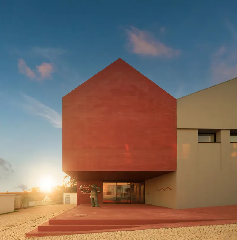 Red tile gable volume raised above the entrance plaza at sunset with two figures