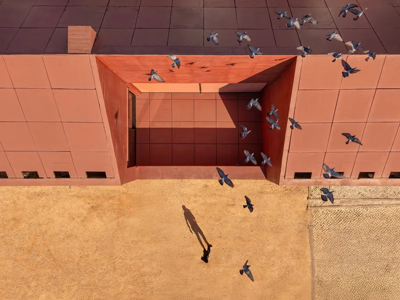 Aerial view of a recessed courtyard in terracotta tile with a single figure and pigeons in flight