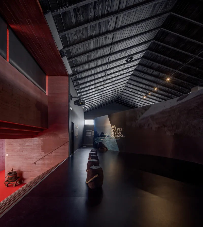 Gallery corridor with exposed timber ceiling beams and a row of wooden barrels under dim lighting