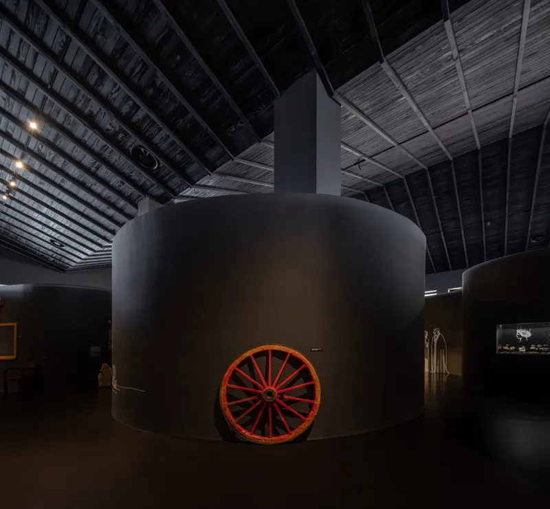 Cylindrical museum display volume with a red wagon wheel at its base beneath an exposed timber ceiling