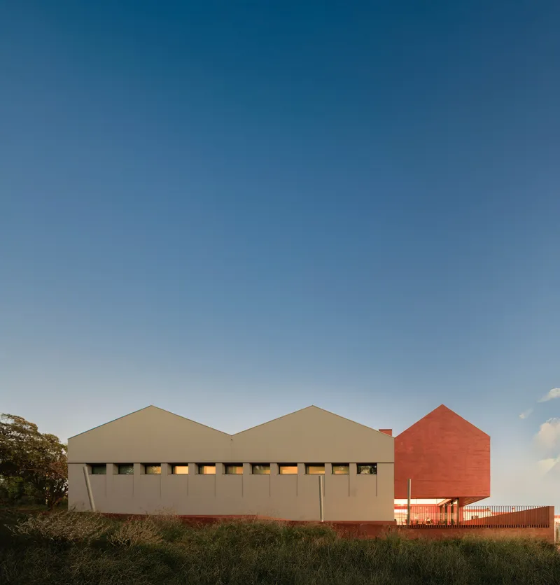 Two gabled volumes clad in beige and red brick alongside a grassy field at dusk
