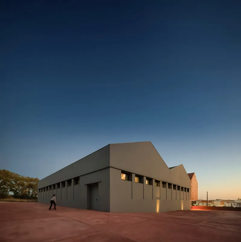 Corner view of the grey paneled facade with square openings and a pedestrian crossing the plaza at twilight