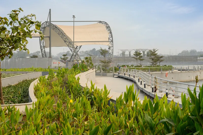 Curved lattice canopy structure shading planted beds with palms and grasses along a waterfront walkway