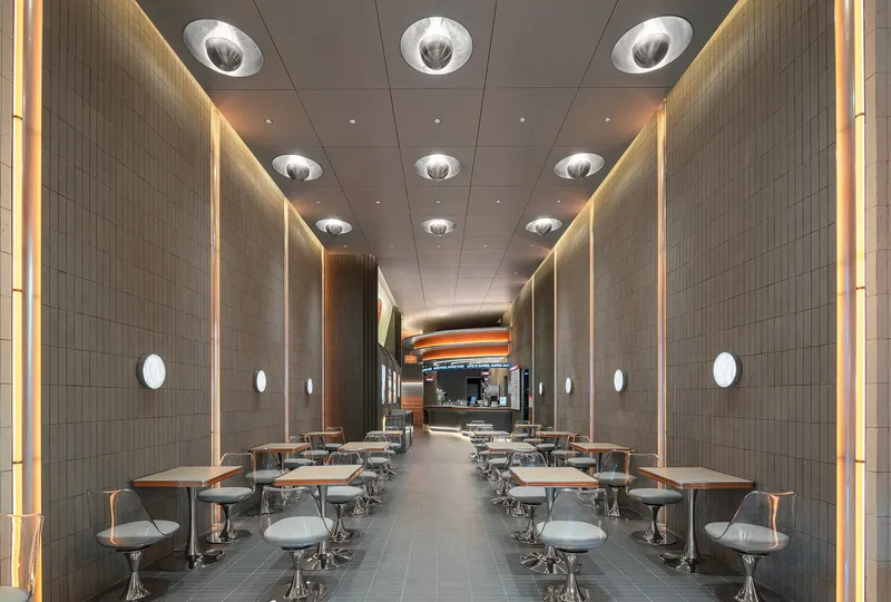 Dining hall with rows of pedestal seating flanking a central aisle beneath recessed circular ceiling lights