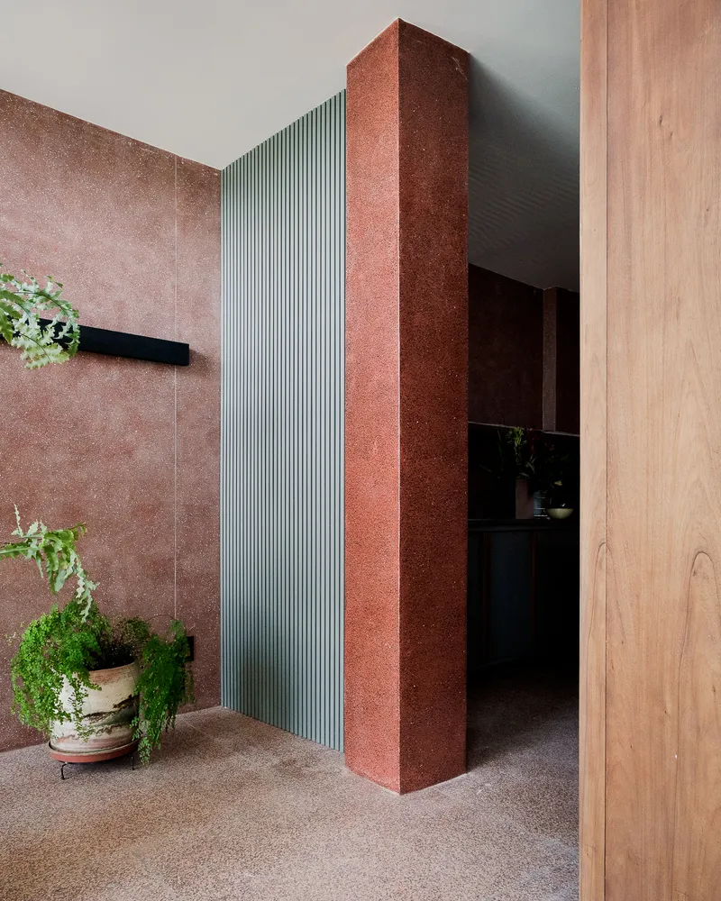 Entrance corridor with red textured stone column, ribbed metal panel and potted ferns on terrazzo floor