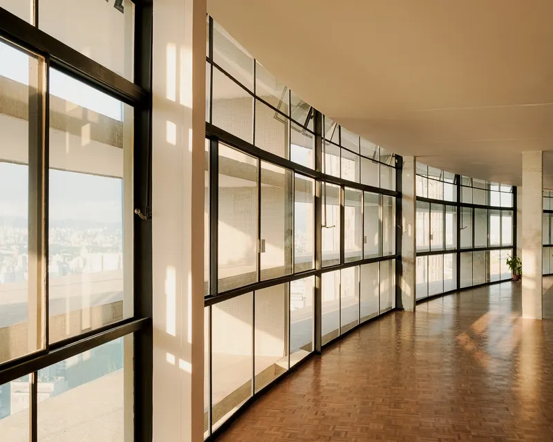 Curved corridor with steel-framed glass partitions and parquet flooring bathed in filtered sunlight