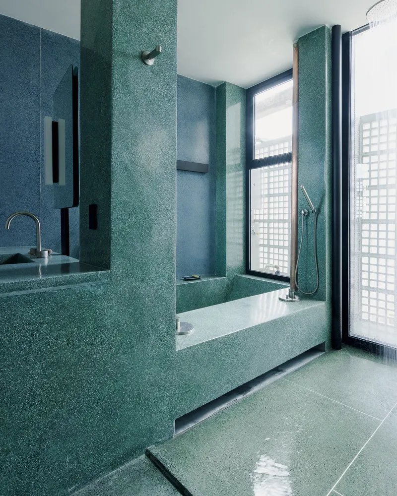 Bathroom with teal terrazzo walls, recessed bathtub and black-framed window opening to a glass brick screen