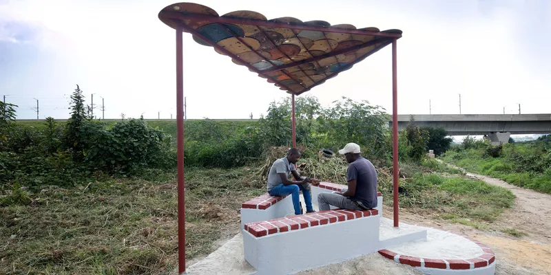 Outdoor seating pavilion with scalloped metal roof canopy and red brick benches in an overgrown field