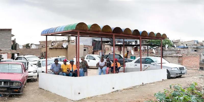 Covered community gathering platform with semicircular roof panels and people seated on white painted benches