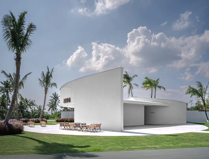 White curved pavilion with cantilevered roof and outdoor terrace surrounded by palm trees and manicured lawn