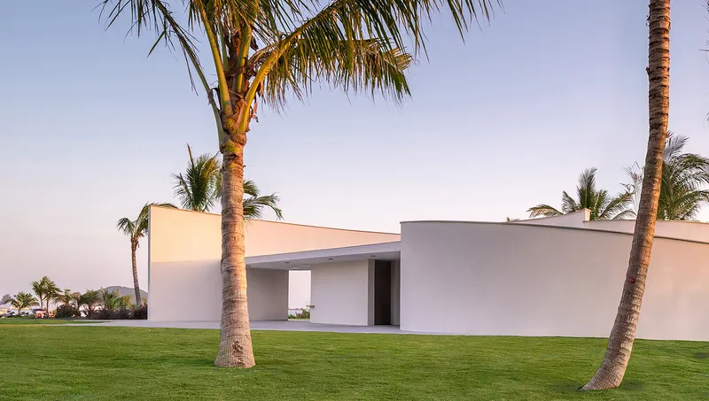 Low horizontal volumes with overhanging roof planes framing openings in a palm grove at dusk