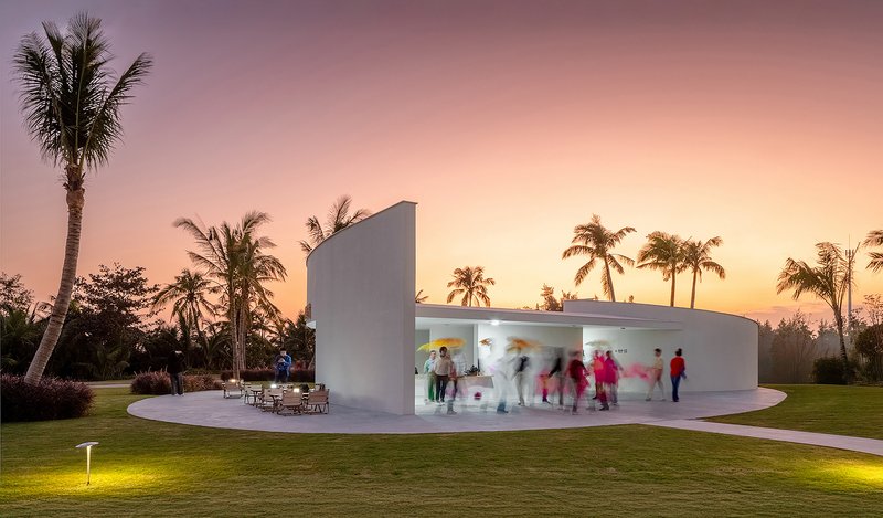 Glazed pavilion with visitors inside at sunset surrounded by palm trees and illuminated lawn