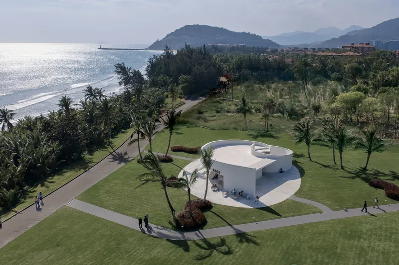 Drone view of the spiral structure with visitors on pathways framed by palms and coastal mountains