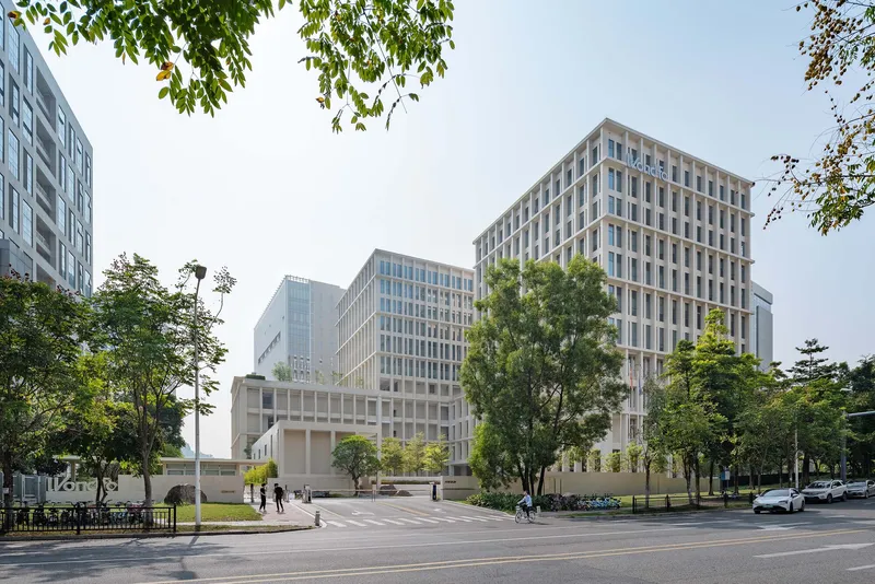 Street view of three gridded office tower volumes framed by mature trees and a pedestrian crossing