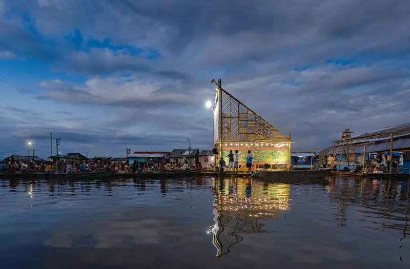 Floating timber pavilion with triangular roof and string lights reflected in calm water at dusk