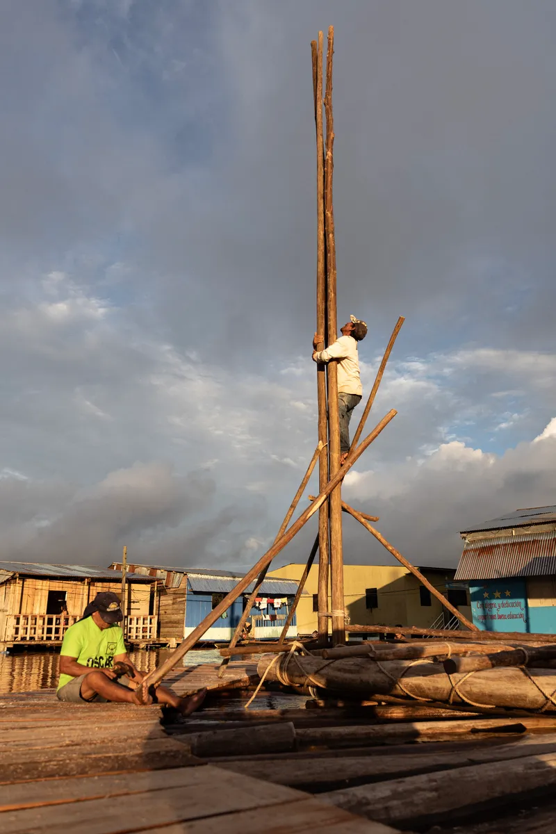 Timber tripod structure being erected on a floating bamboo platform at golden hour