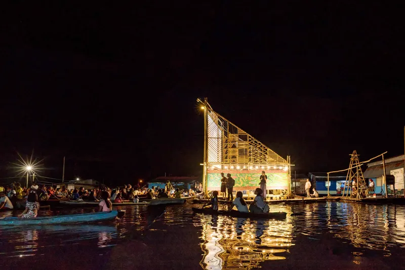 Illuminated bamboo lattice screen and projection wall reflected in dark water with boats nearby at night