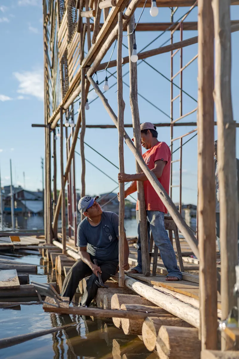 Two workers fitting timber members into a bamboo pole framework under afternoon sun