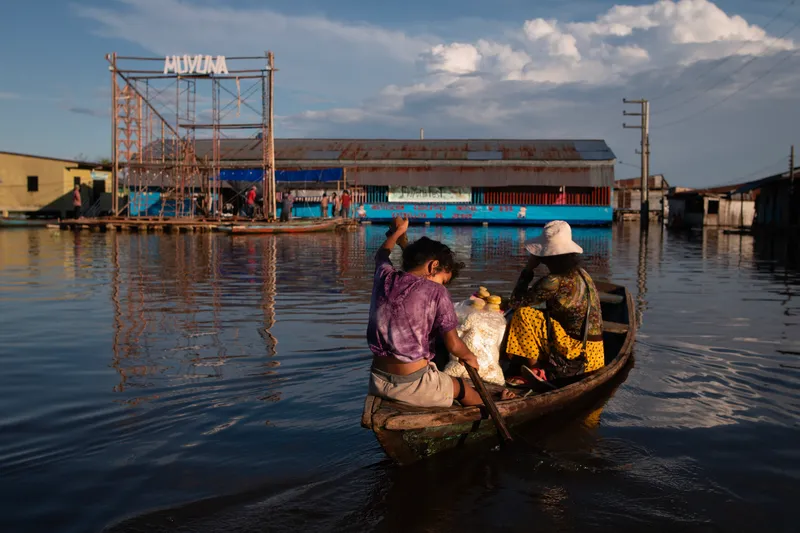 Floating blue school structure viewed from a wooden boat approaching across flooded waters in late afternoon