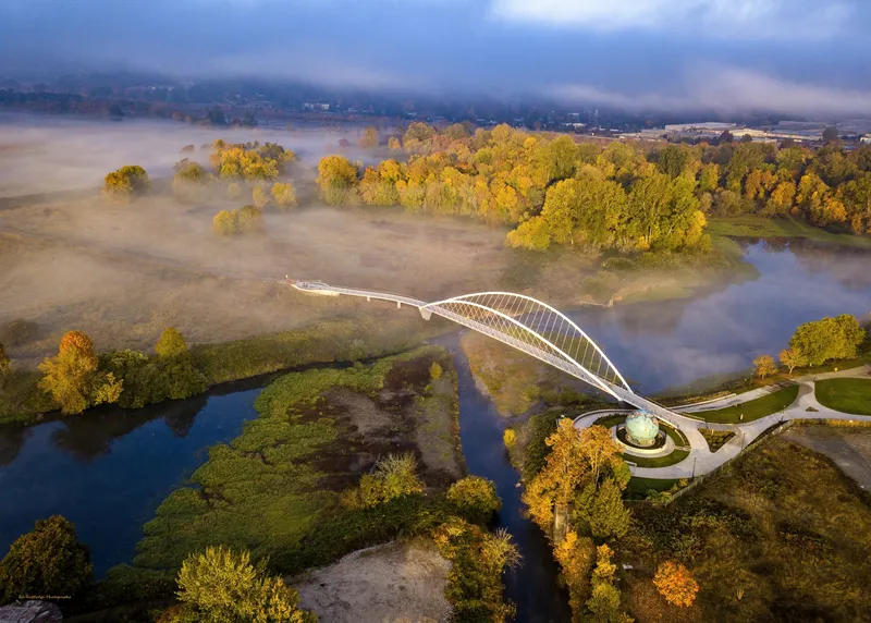 Aerial view of the white arched pedestrian bridge spanning the river amid autumn trees and morning fog