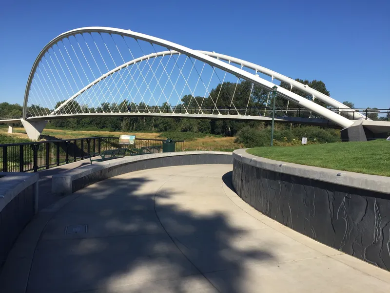Curved concrete plaza with textured retaining walls leading toward the white arched bridge