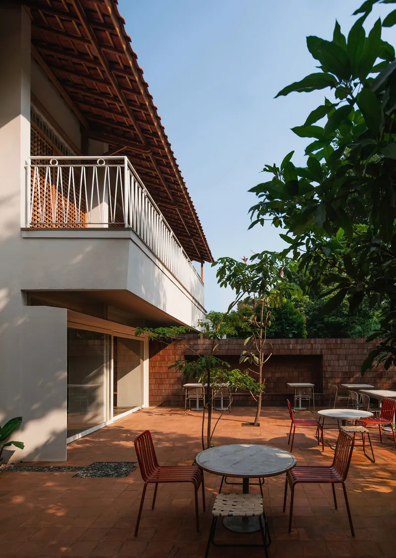Courtyard corner showing upper balcony with clay tile overhang and trees beside outdoor seating