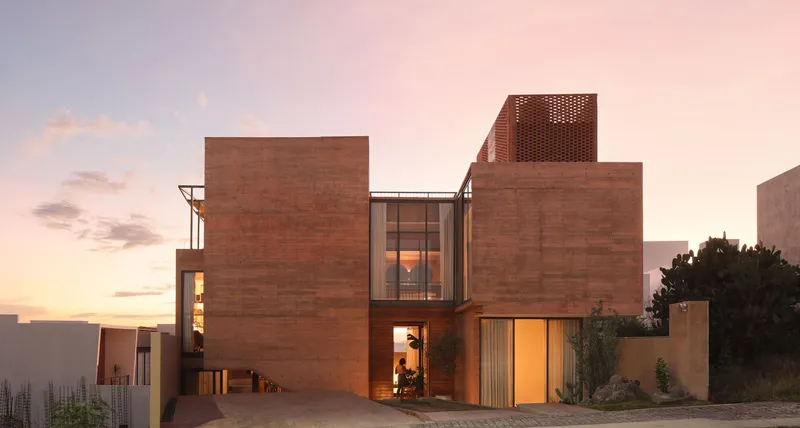 Rammed earth facade with stacked volumes and perforated brick tower at dusk