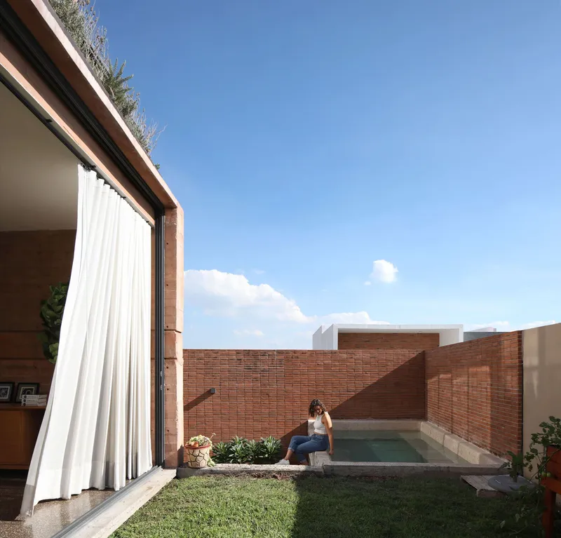 Private courtyard with brick walls, lawn, pool and person seated beside a planted bed under blue sky