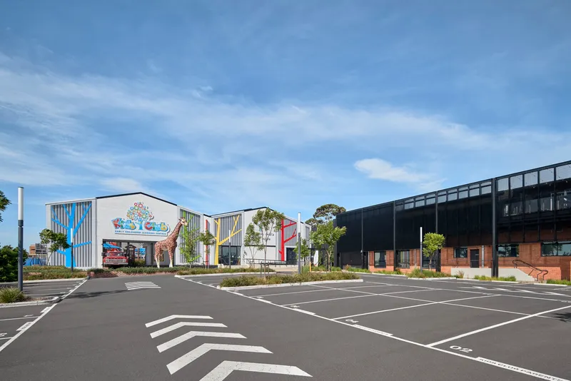 Parking lot view of gabled building cluster with corrugated metal cladding and coloured facade graphics under clear sky