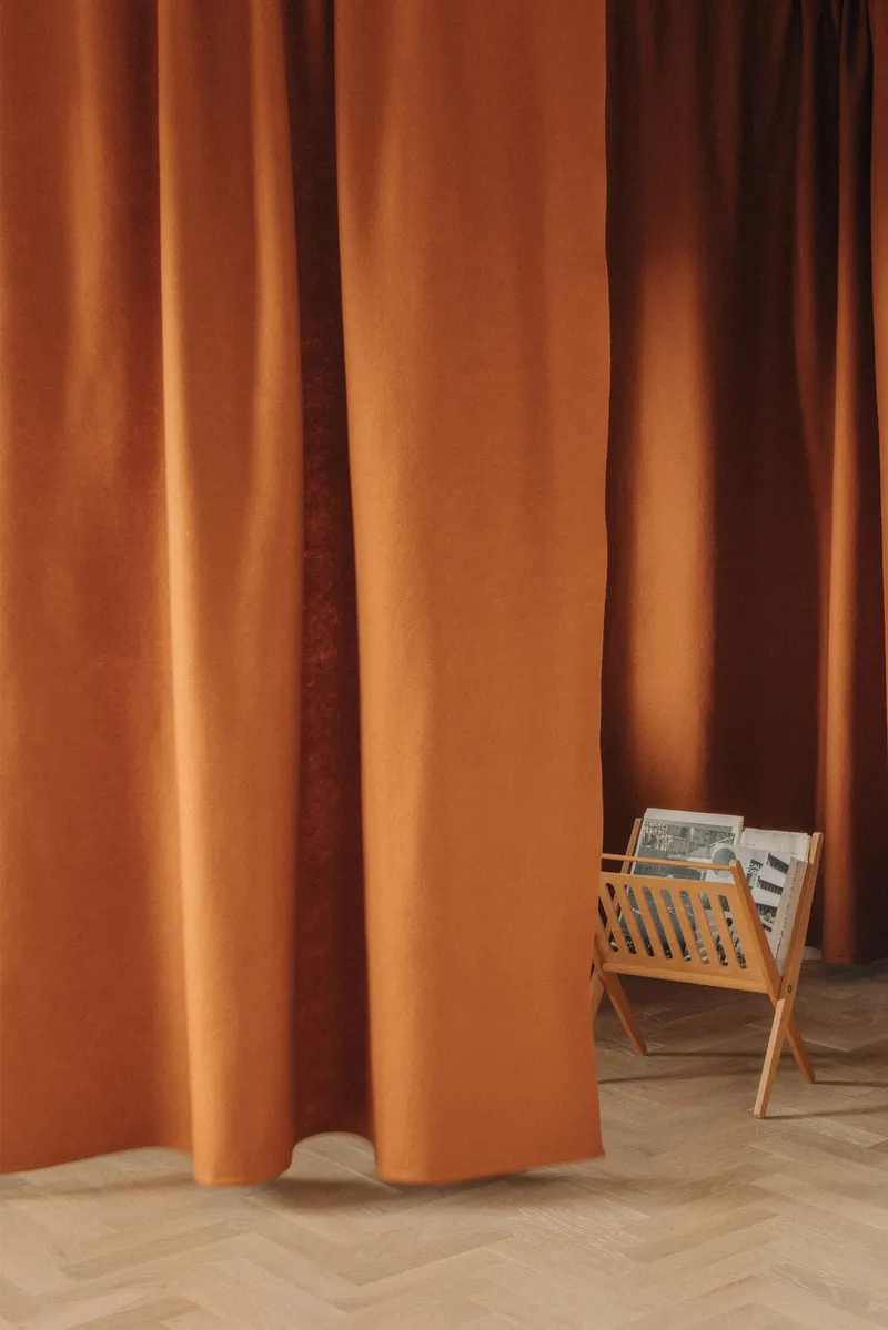 Floor-to-ceiling terracotta curtains draping over herringbone timber flooring beside a timber magazine rack