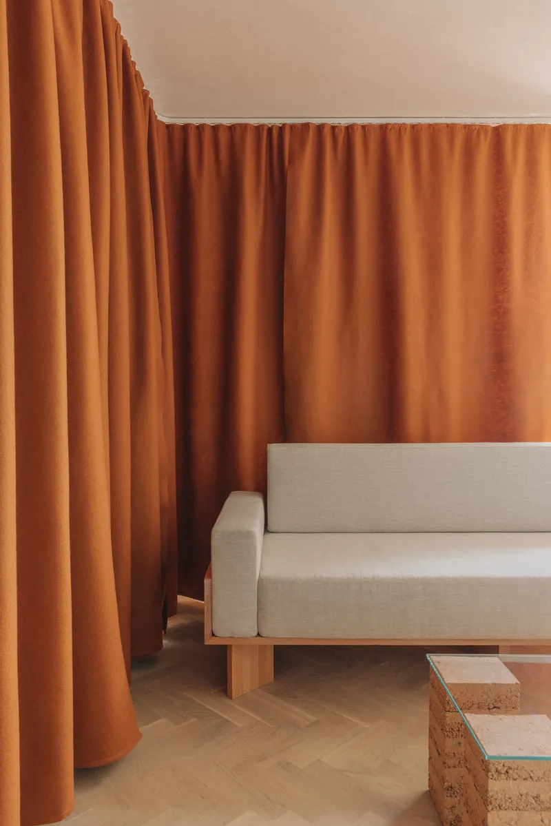Upholstered seating on timber plinth beside cork and glass side tables framed by terracotta curtains
