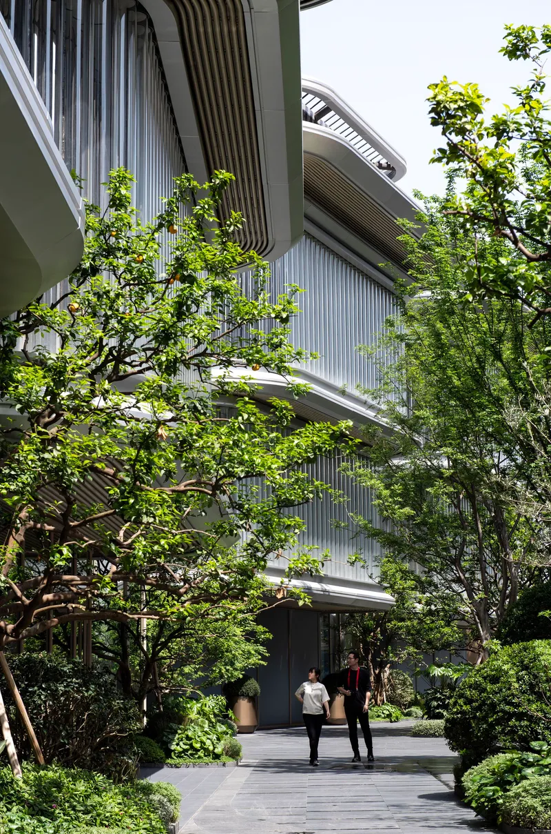 Ground-level entry path with two figures walking beneath louvered balconies and surrounding foliage