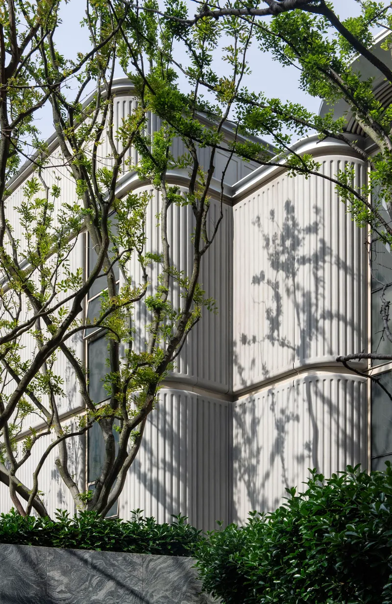 White vertically ribbed facade with curved corners casting dappled shadows from overhanging tree branches