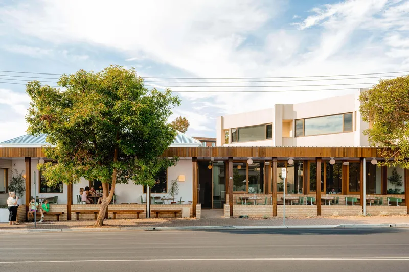 Street facade with timber-framed glazing, white upper volume, and established trees shading the outdoor seating area