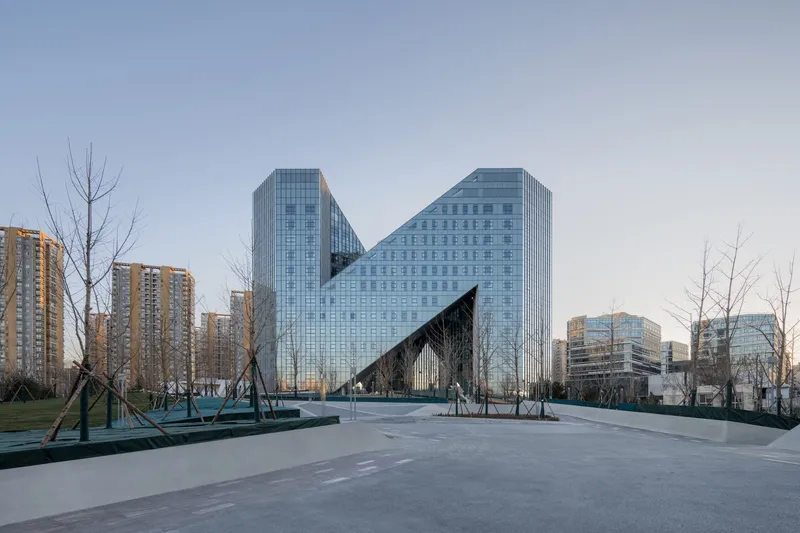 Zigzag glass curtain wall facade viewed from a paved plaza with bare winter trees