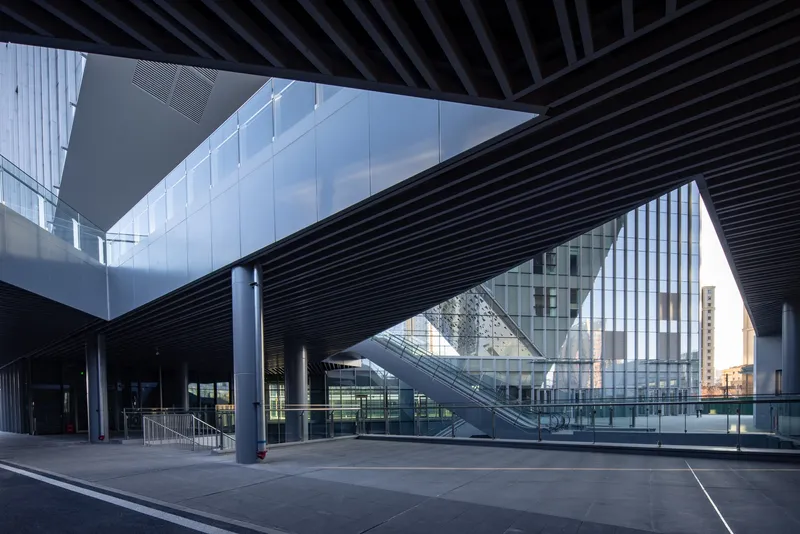 Covered plaza beneath black slatted canopy with escalator and glass facades reflecting twilight sky