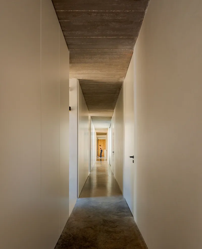 Narrow corridor with board-formed concrete ceiling and white walls leading to a figure at the end