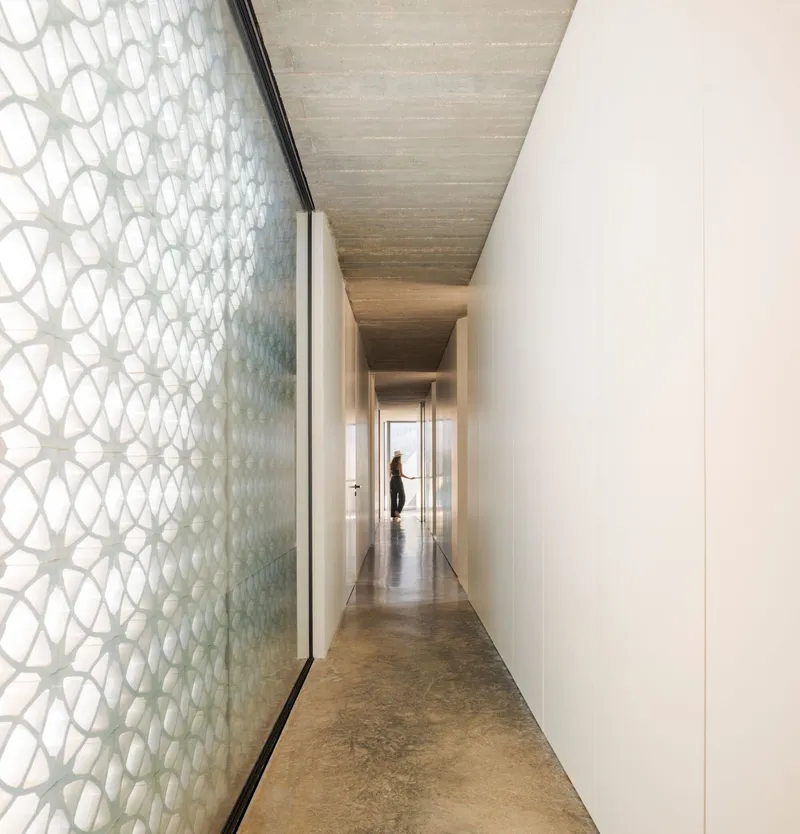 Corridor with patterned glass screen wall and timber ceiling viewed toward a figure in daylight
