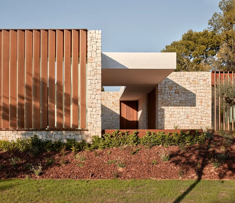 Street facade with vertical corten steel panels, stone masonry, and white stucco volumes amid planted beds
