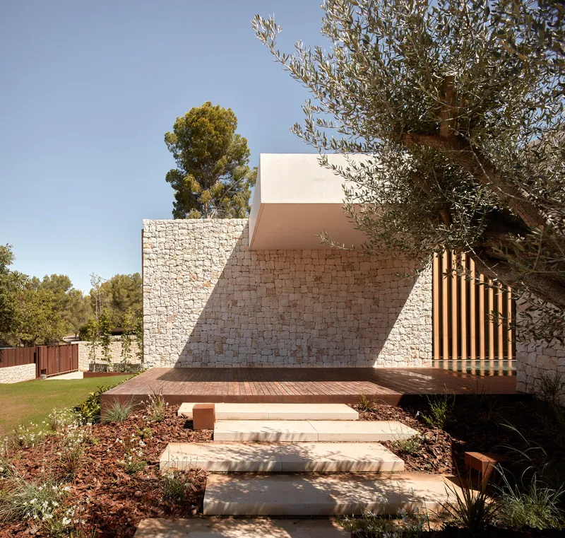 Exterior stairway with floating concrete treads leading to timber deck beside angled stone wall and olive tree