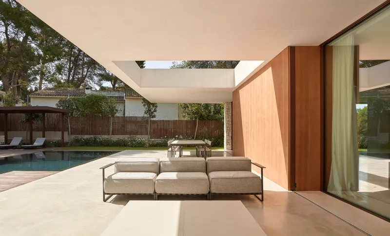 Covered outdoor terrace with wood-paneled pivot door and low sofa overlooking a pool and timber fence