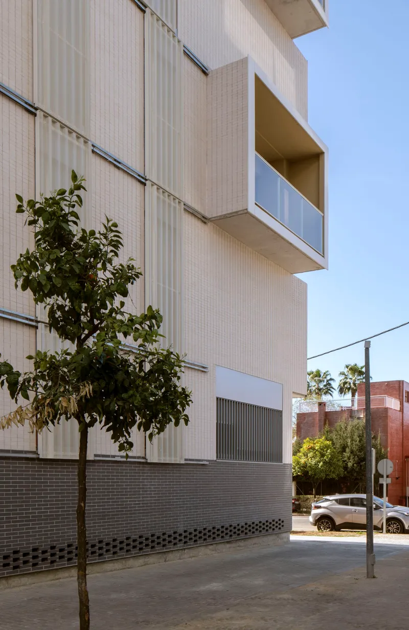 Close-up of the facade with white ribbed panels, recessed balcony, and street tree in sunlight
