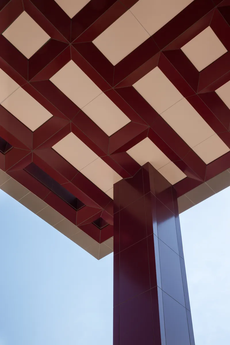 Detail of the red metal pergola with woven beam pattern meeting a column against pale blue sky