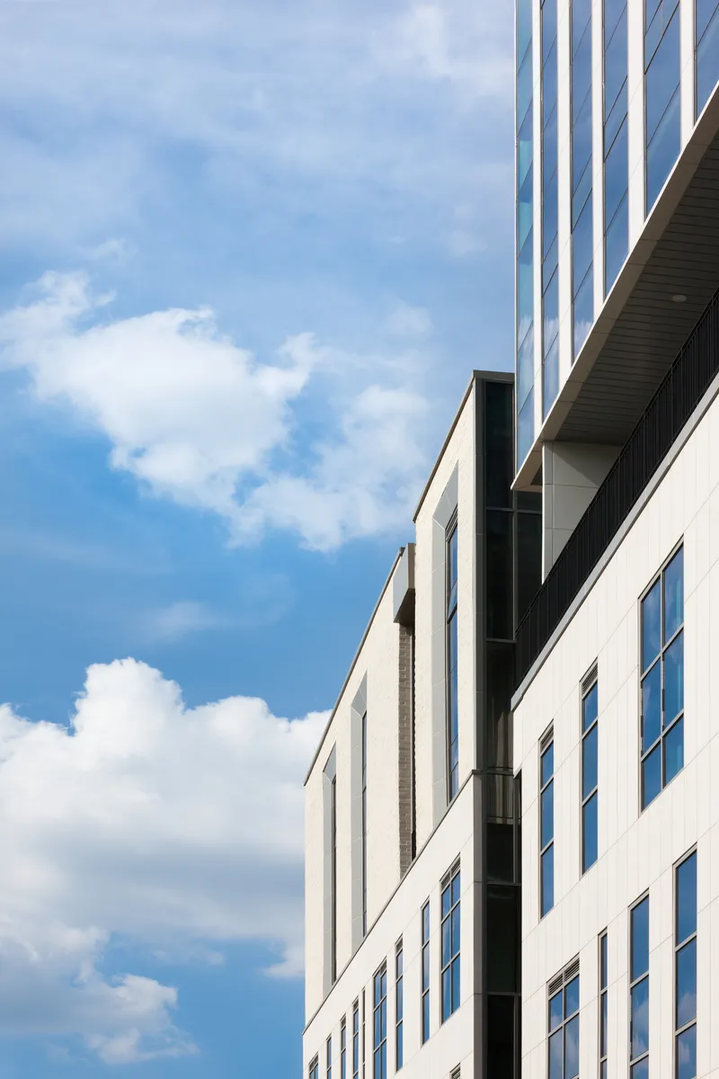 Oblique view of white masonry facade meeting glazed tower volume under scattered afternoon clouds