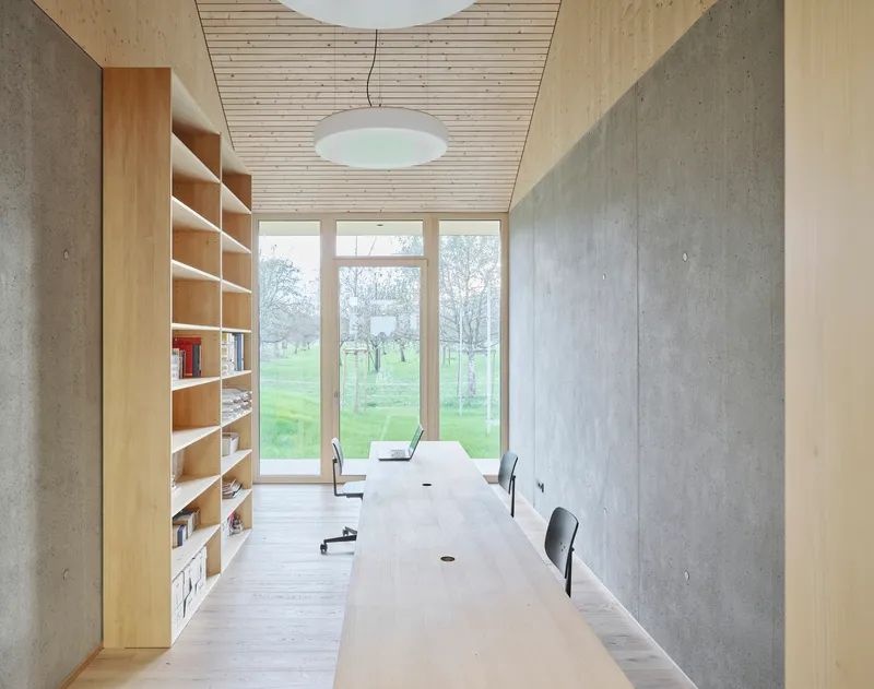 Long interior workroom with plywood shelving, concrete wall, timber table and disc pendant above