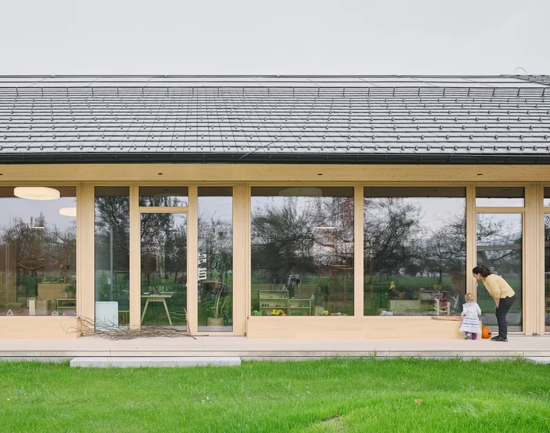 Glazed facade with timber mullions beneath roof overhang showing figure bending near entrance